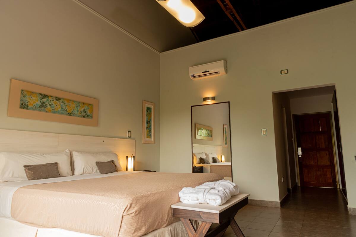 A hotel room with a large bed, a bedside table with towels, a wall-mounted air conditioner, and a door leading to another room.