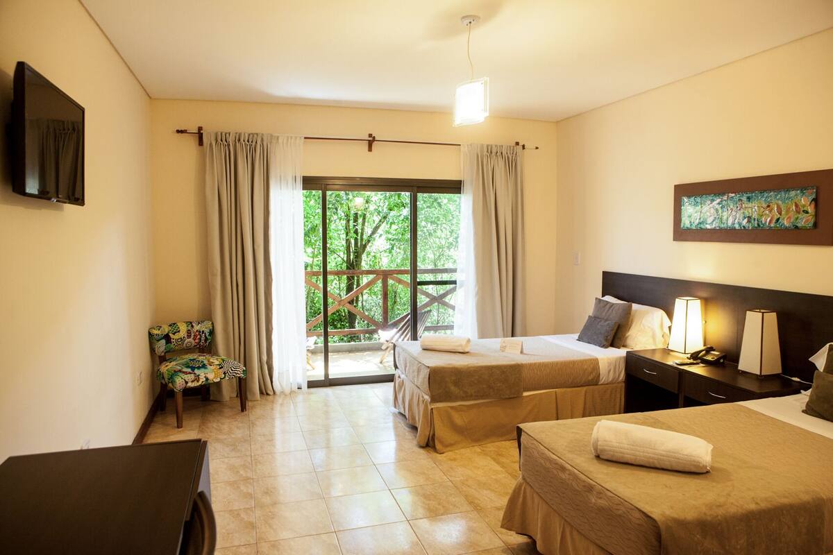A hotel room with two beds, a TV, a desk, and a balcony with a wooden railing.