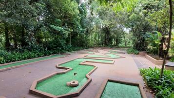 A miniature golf course with green artificial turf and numbered holes, surrounded by dense foliage.