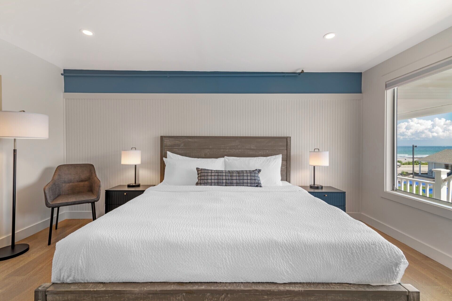 A bedroom with a large bed, two bedside tables with lamps, a chair, and a view of the beach.