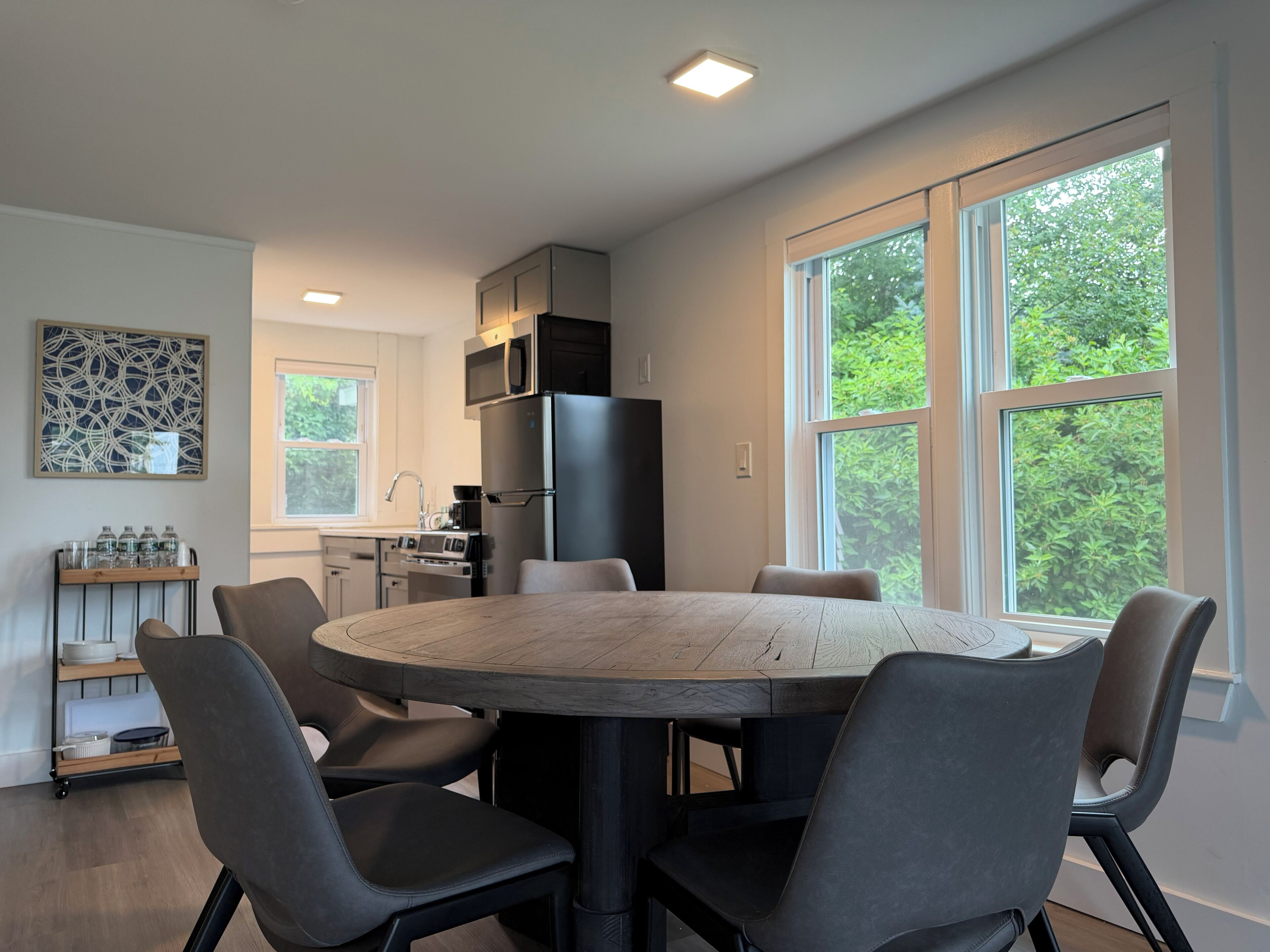 A modern kitchen with a central island, a dining table with chairs, and large windows with a view of greenery.