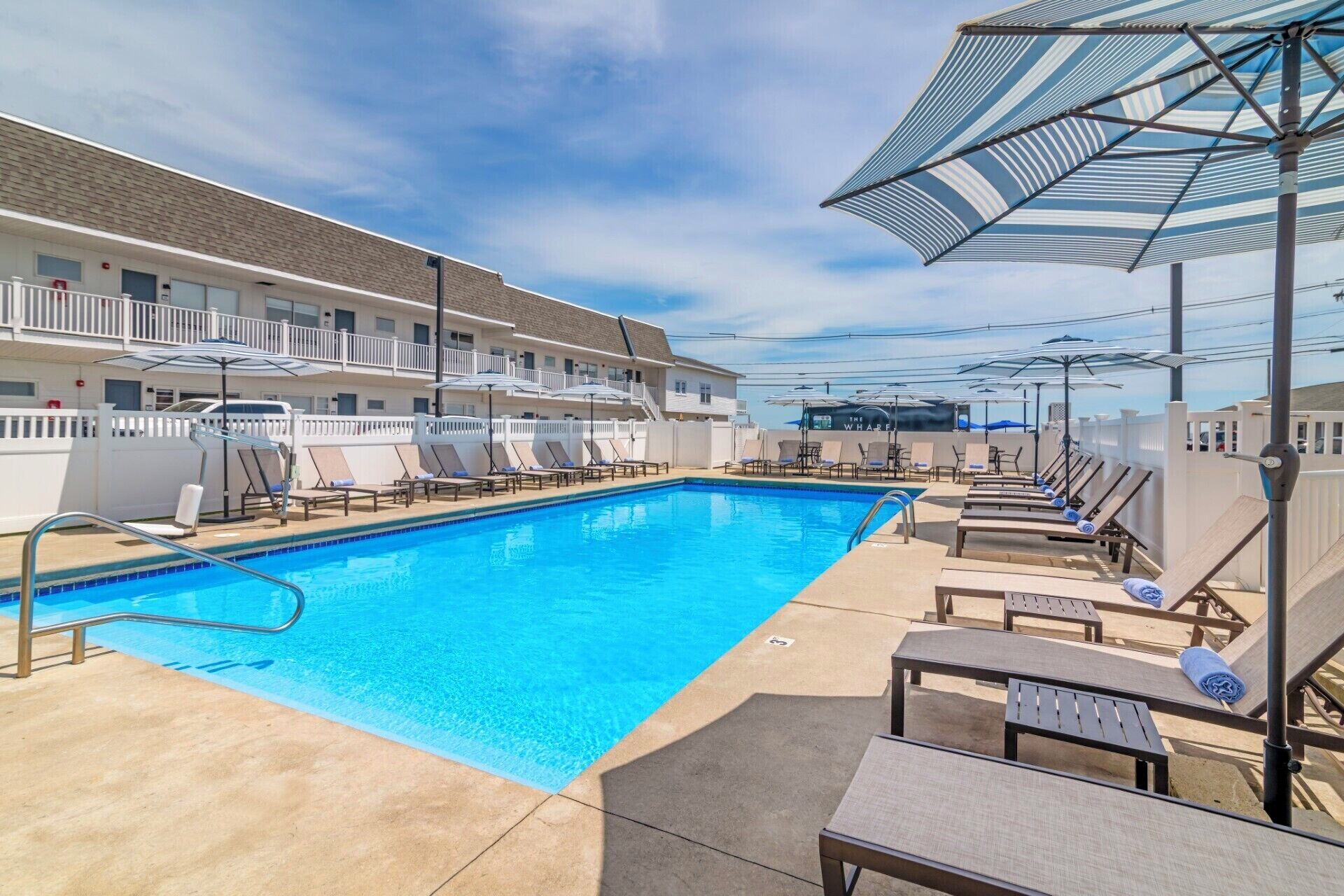A swimming pool with lounge chairs and umbrellas, a white fence, and a building with balconies.