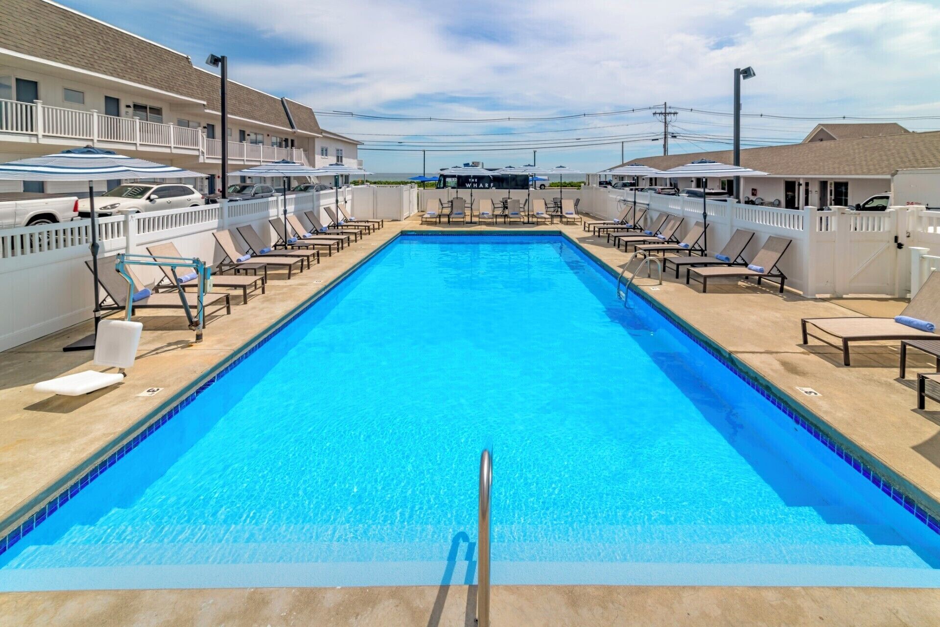 A rectangular swimming pool with lounge chairs and umbrellas on both sides, surrounded by a white fence.