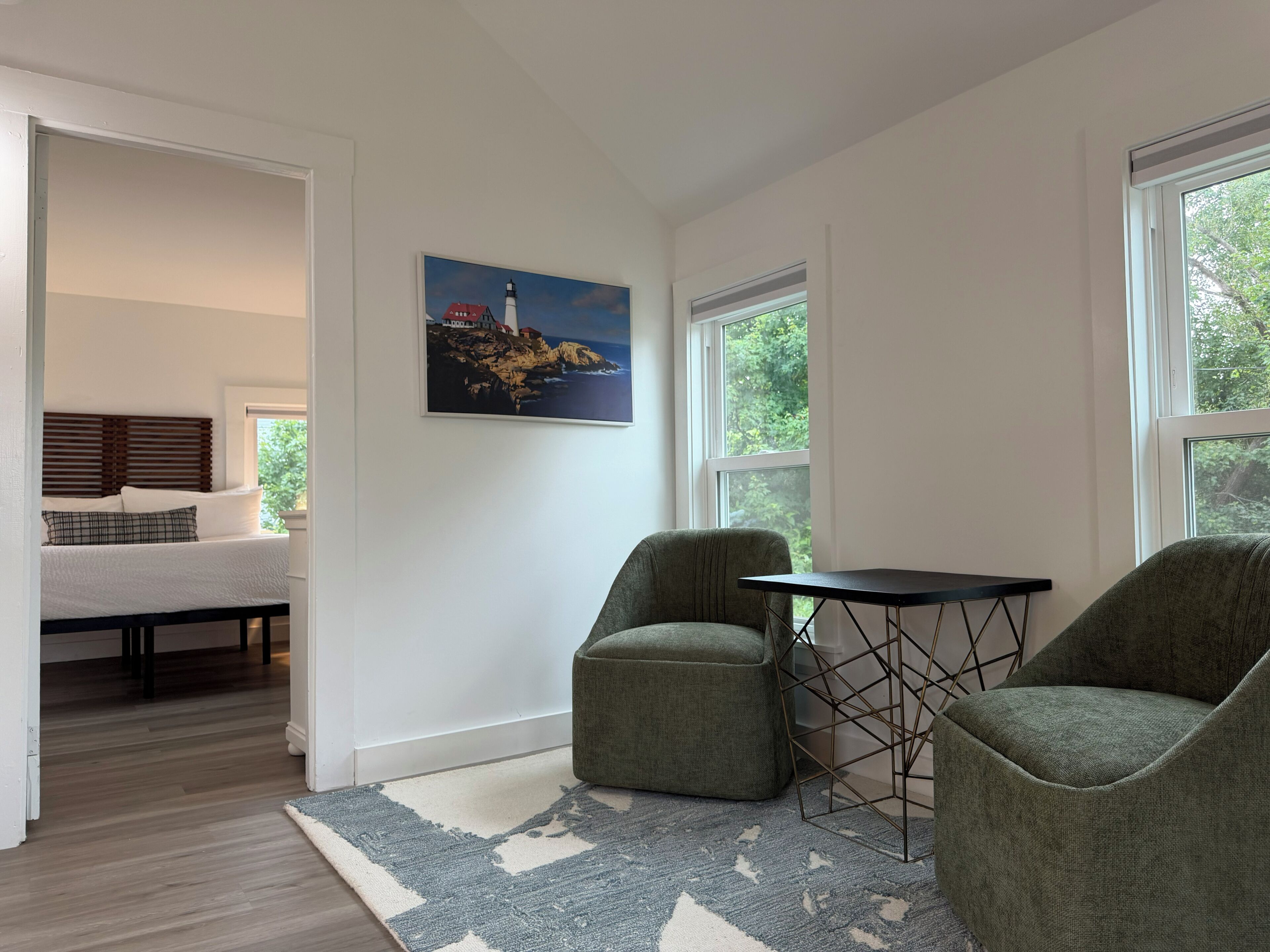 Ein modernes Zimmer mit einem Bett, einem Gemälde, zwei Stühlen, einem Tisch und großen Fenstern.