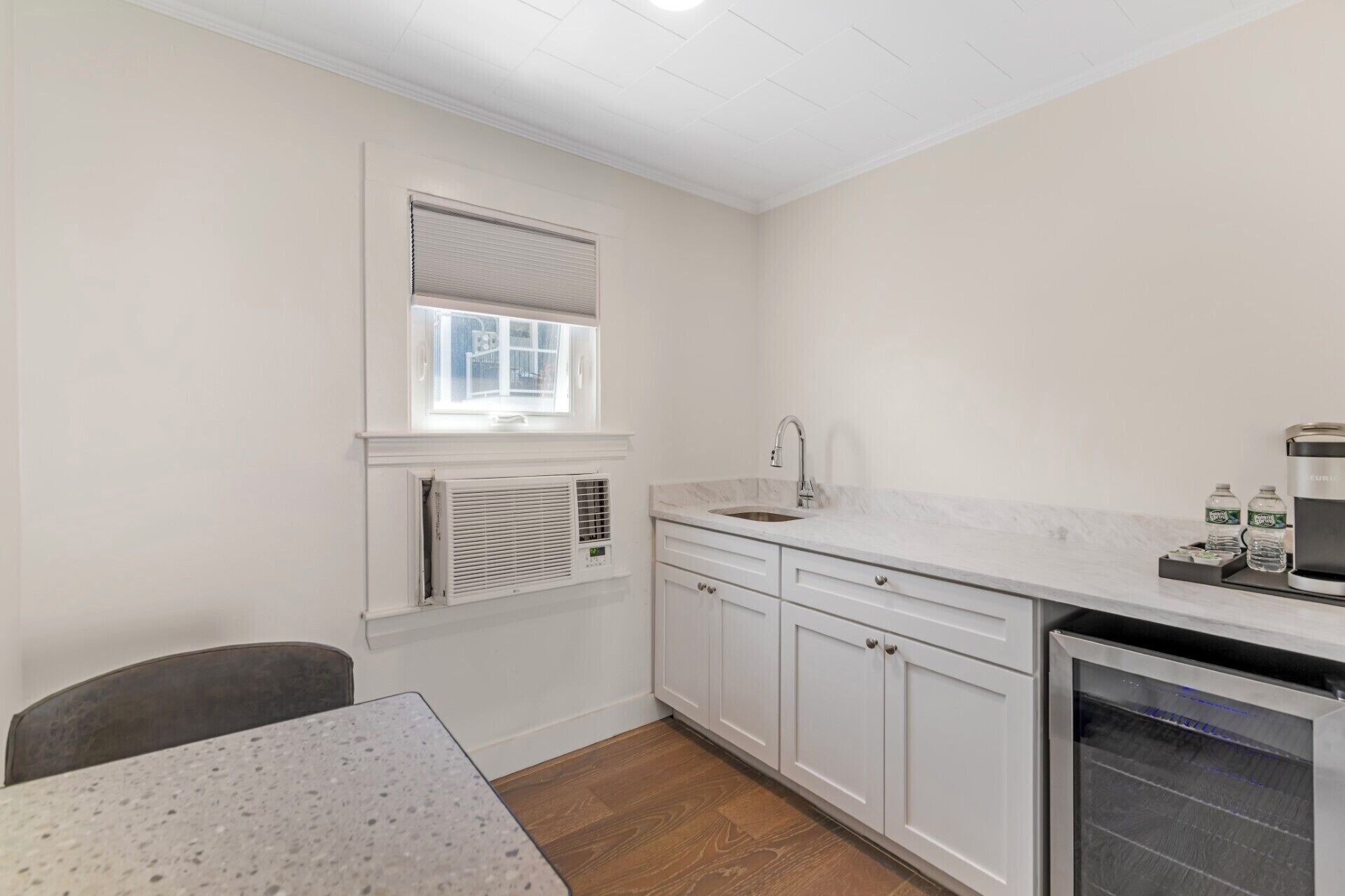 A compact kitchen with white cabinets, a sink, a window with blinds, a built-in air conditioner, and a refrigerator.