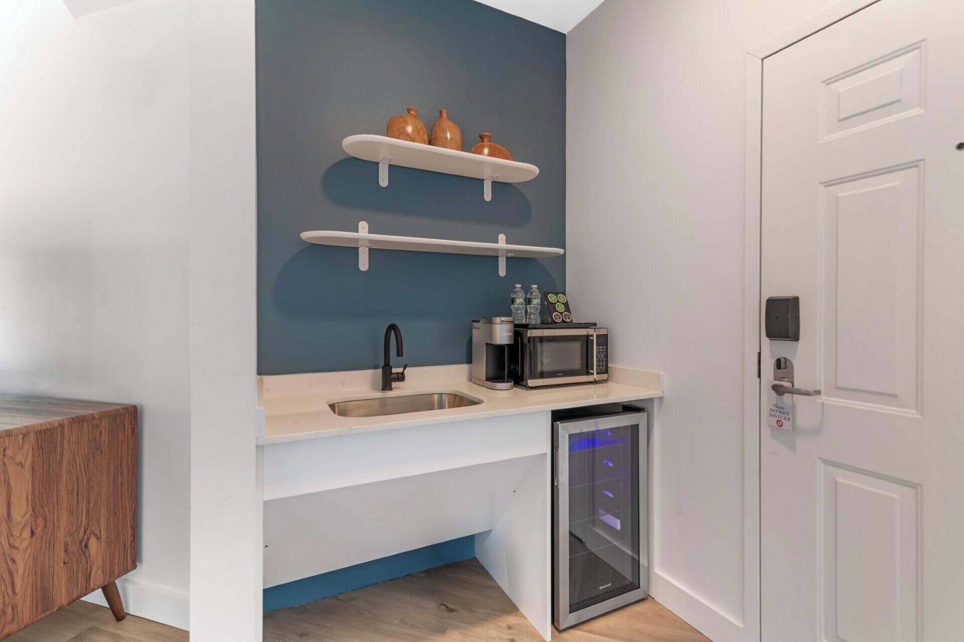 A compact kitchen with a sink, microwave, and coffee maker. There are two shelves above the sink, and a door with a 'Do Not Disturb' sign.