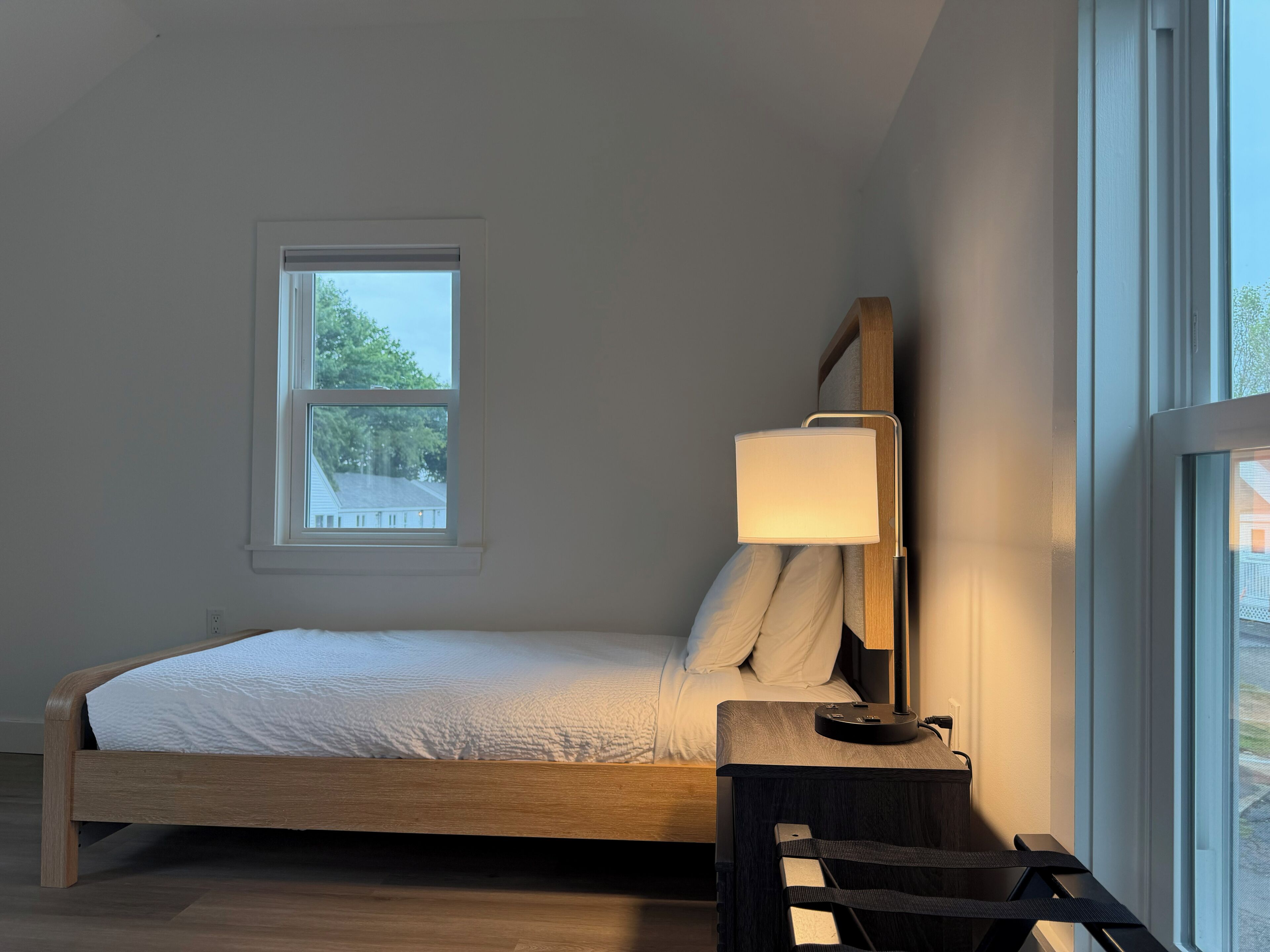 A bedroom with a bed, a bedside table with a lamp, a window, and a view of trees.