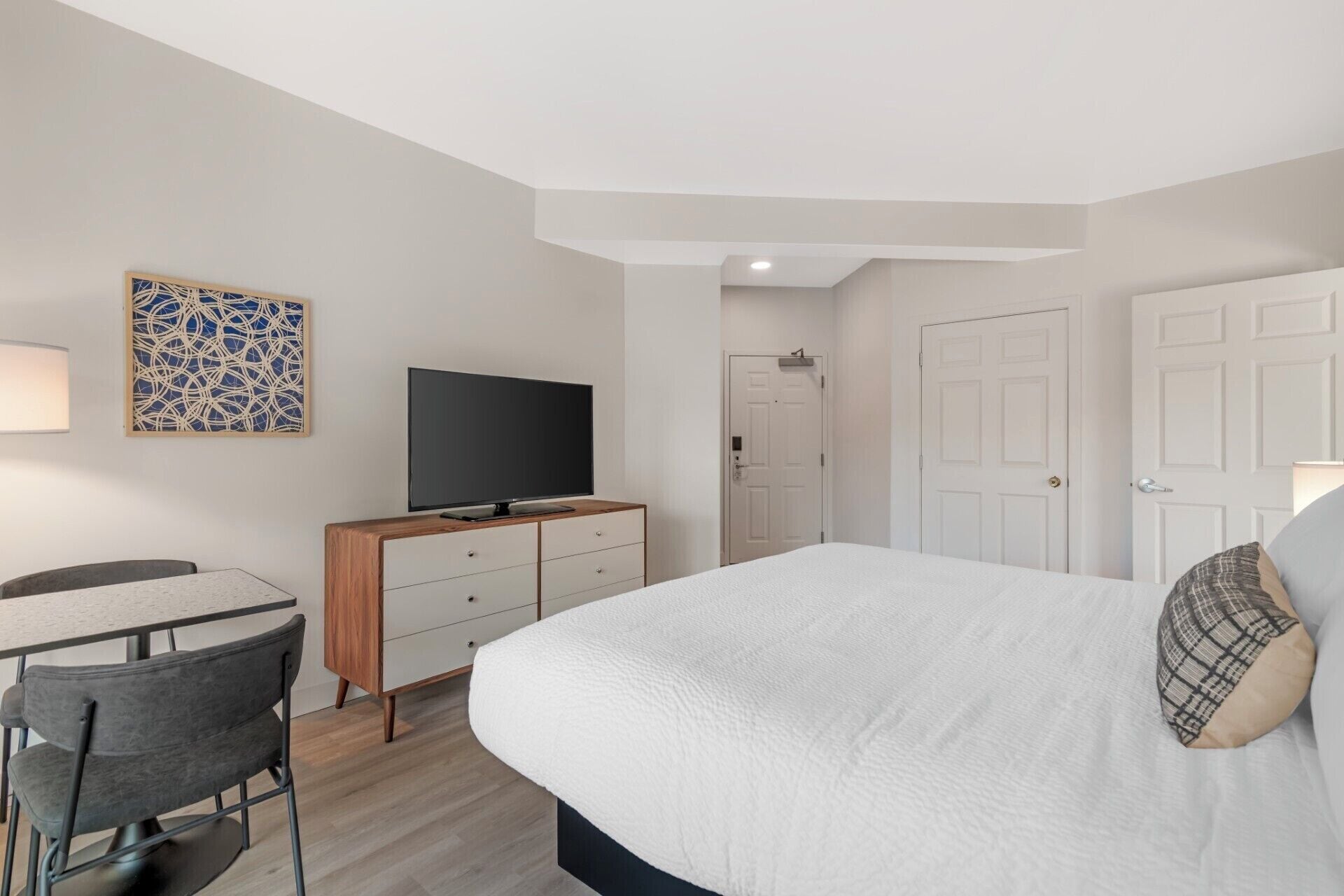 A hotel room with a bed, a desk with a chair, a television, a door, and a wall decoration.