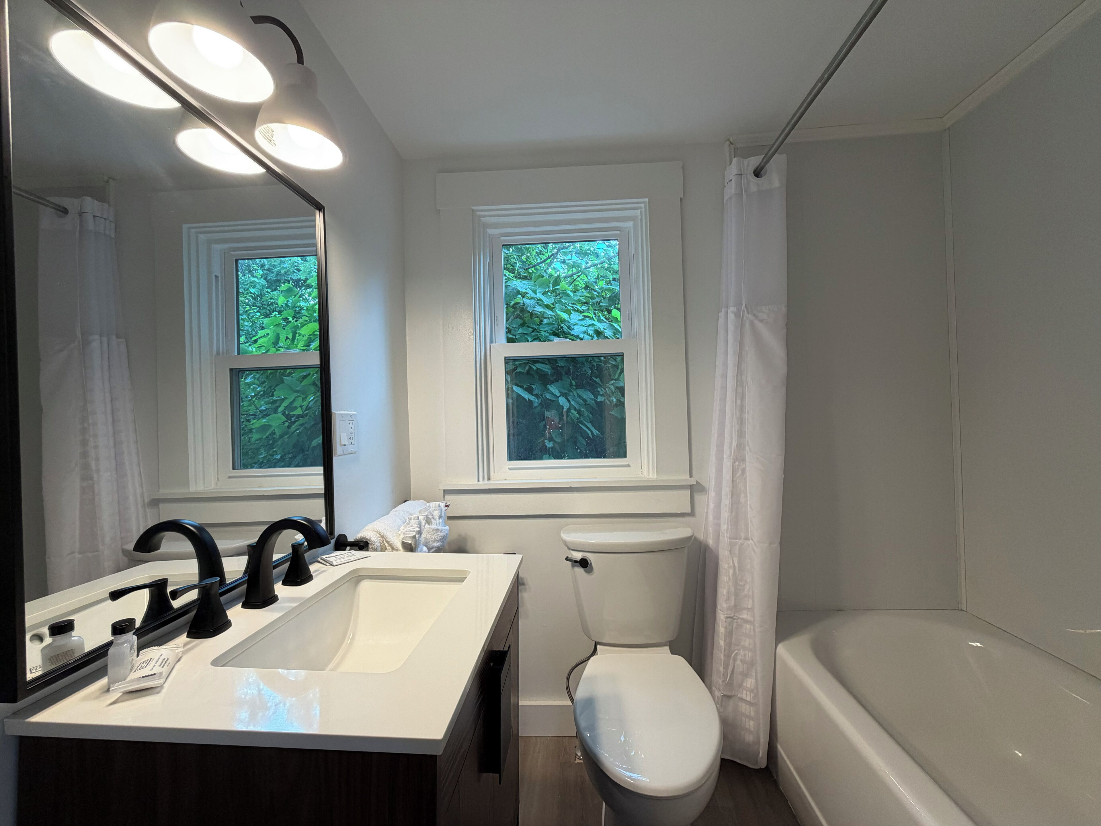 Un baño con un lavamanos blanco, un grifo negro, un inodoro y una bañera. Hay dos ventanas con marcos blancos y vistas a la vegetación exterior.