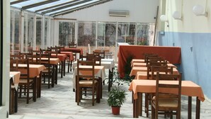 Un área de comedor al aire libre con techo, varias mesas y sillas, una planta en maceta y vistas al exterior.
