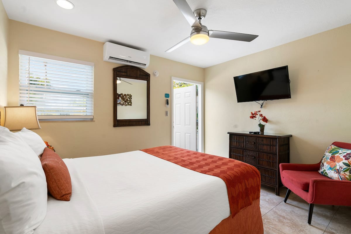 A hotel room with a bed, a ceiling fan, an air conditioning unit, a television mounted on the wall, a dresser, and a red armchair.