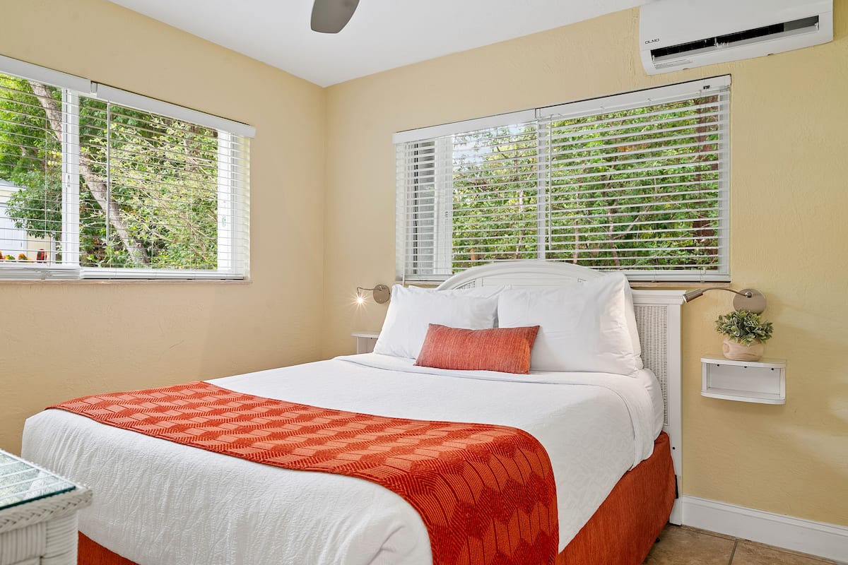 A neatly made bed with white and orange bedding, two bedside tables with lamps, and a window with blinds.