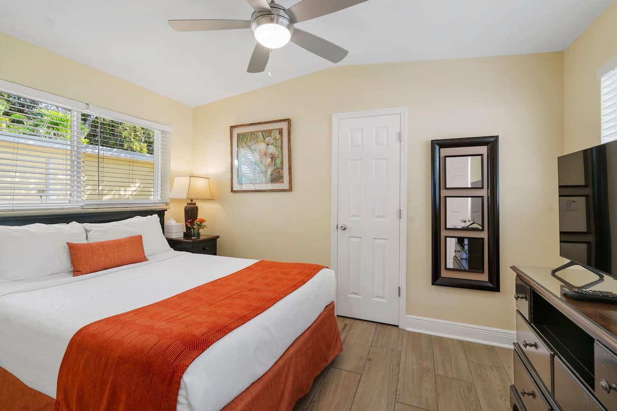 A hotel room with a bed, a ceiling fan, a door, a framed picture, a dresser, and a television.