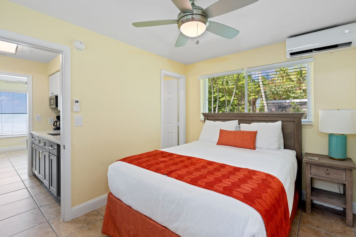 A hotel room with a bed, a nightstand, a ceiling fan, an air conditioning unit, a window with a view of trees, and a kitchenette with cabinets and a microwave.