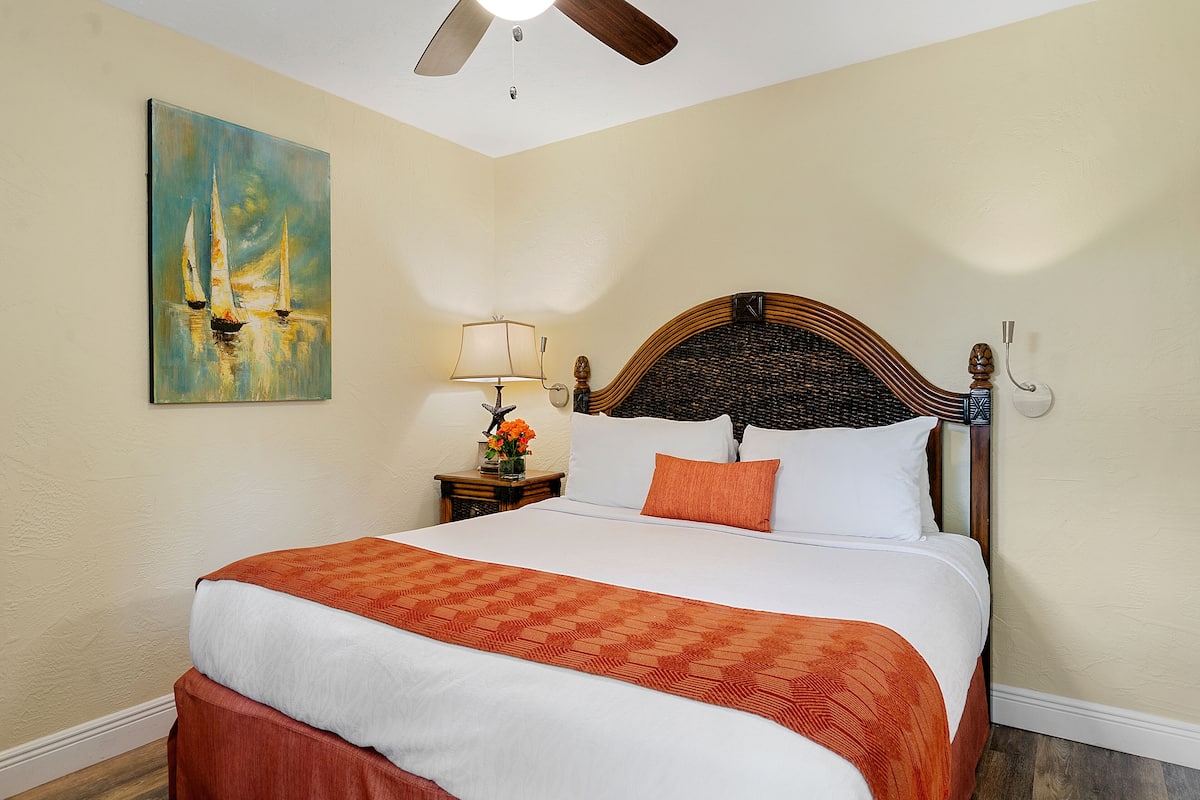 A neatly made bed with a patterned bedspread, a wooden headboard, and a nightstand with a lamp and vase of flowers.