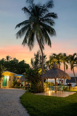 A tropical resort with a thatched-roof hut, palm trees, and a view of the ocean at sunset.