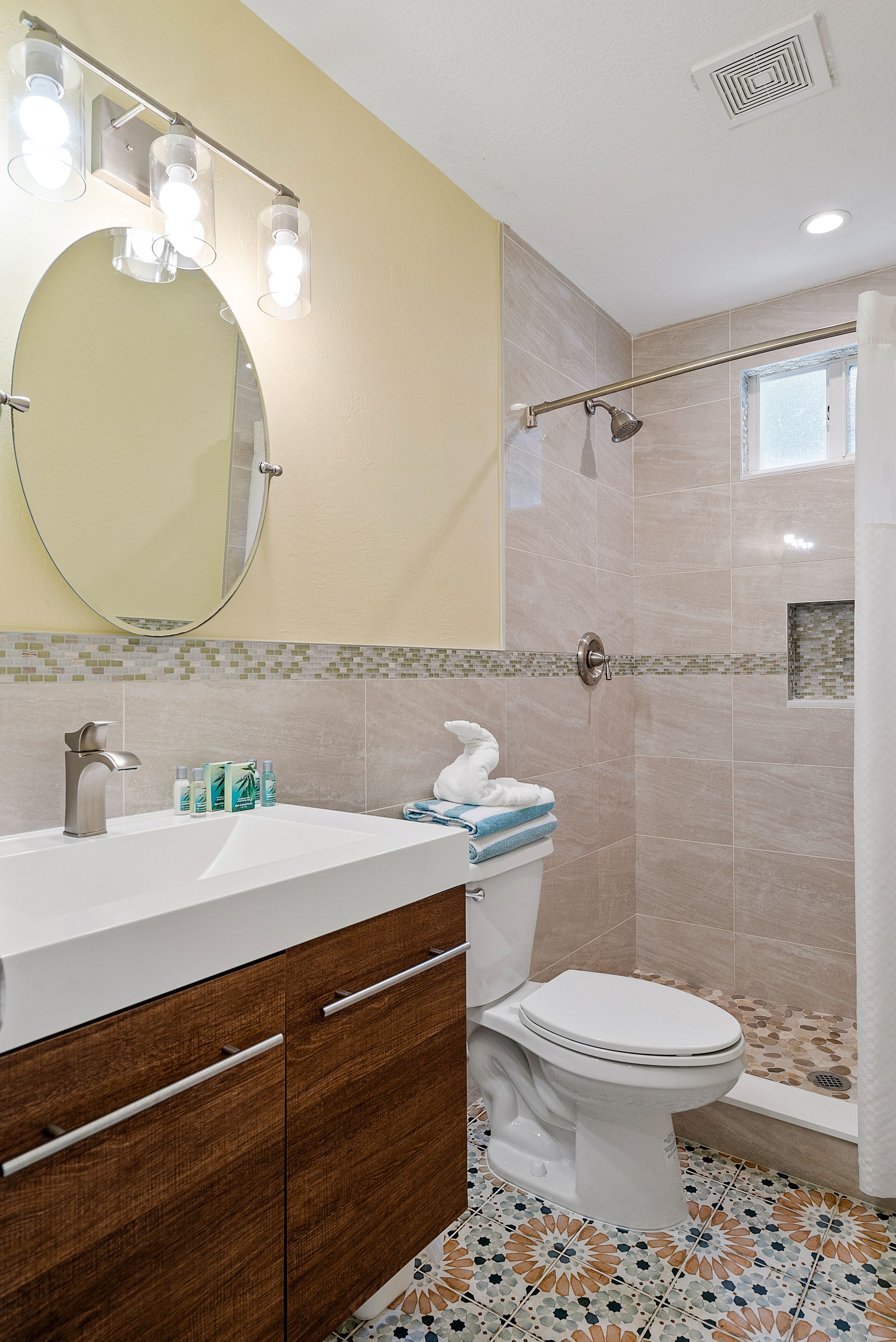 Un baño con lavamanos blanco, espejo, inodoro, zona de ducha y baldosas de suelo estampadas.