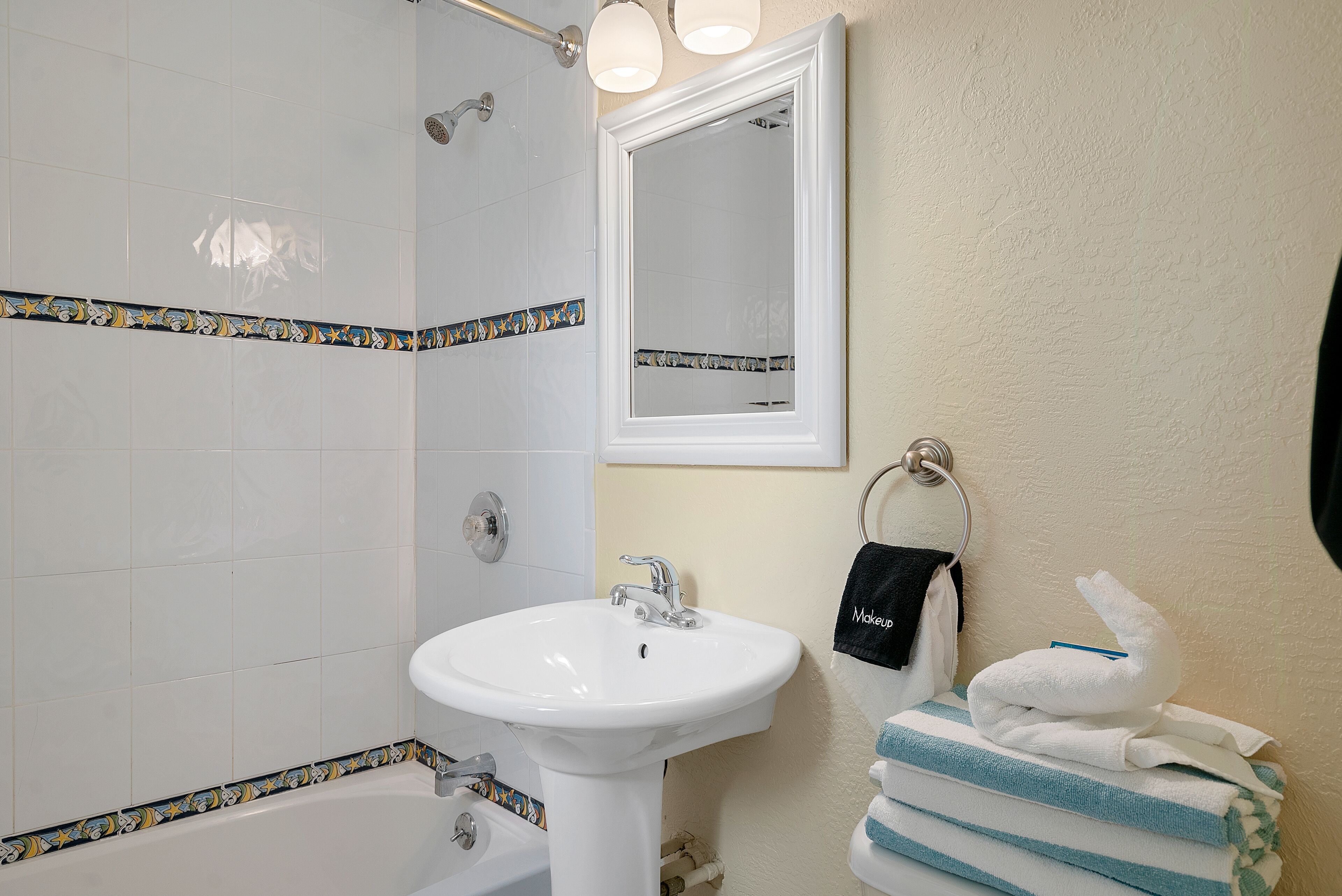 Un baño con un lavamanos blanco, una bañera y una ducha. Hay un perchero con toallas y una toalla negra con la palabra "Makeup" escrita.