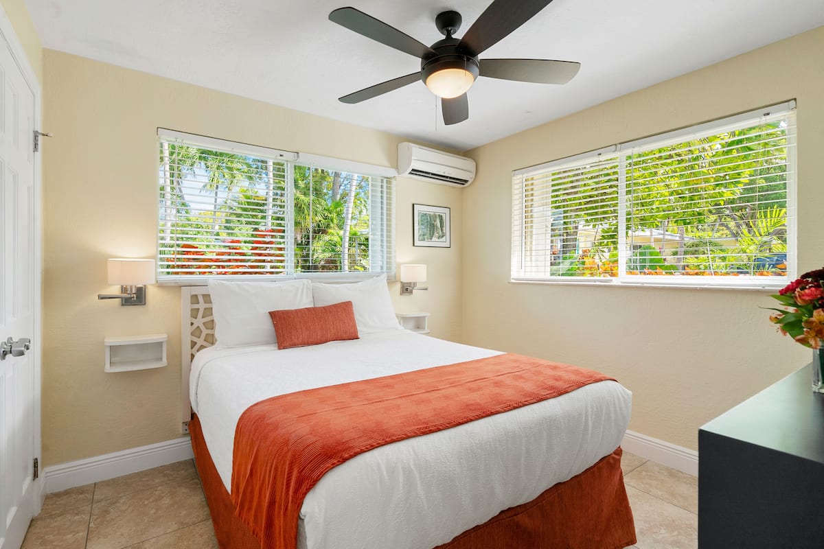 A hotel room with a bed, a ceiling fan, two windows with blinds, an air conditioning unit, and a nightstand with a lamp.