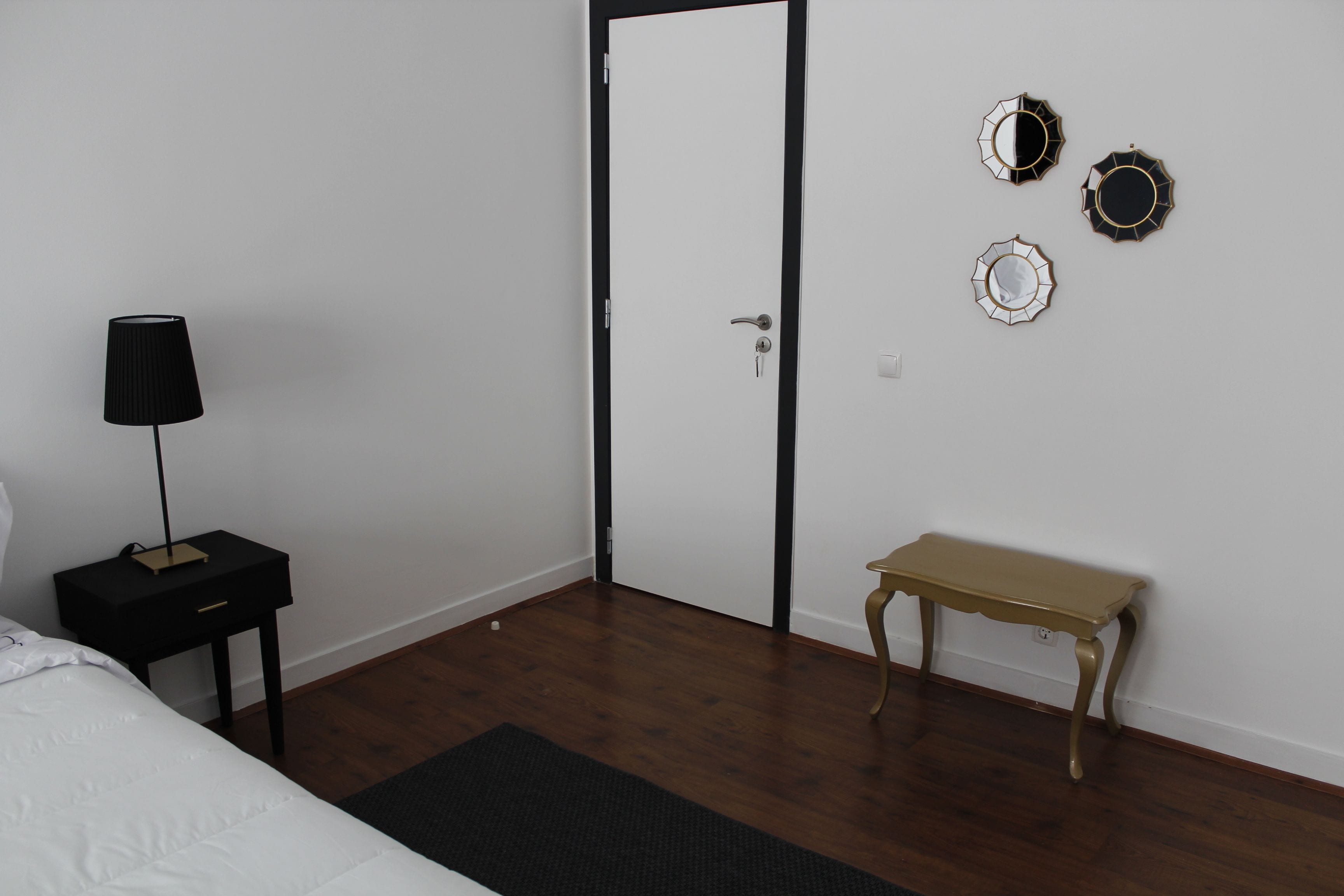 Een slaapkamer met een bed, een nachtkastje met een lamp, een houten bankje en een deur.