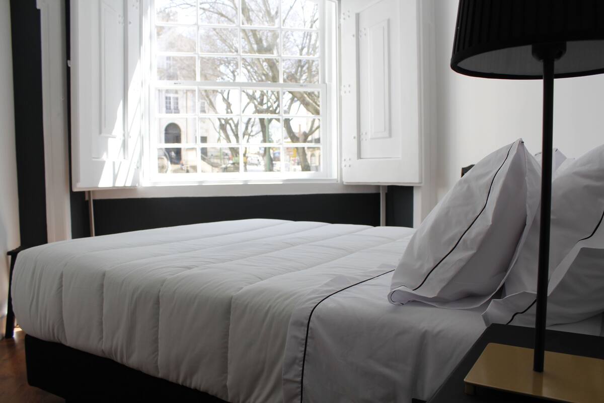 A neatly made bed with white bedding, a bedside table with a lamp, and a window with a view of trees and buildings.