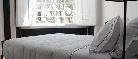 Una cama bien hecha con ropa de cama blanca, una mesita de noche con una lámpara y una ventana con vistas a árboles y edificios.