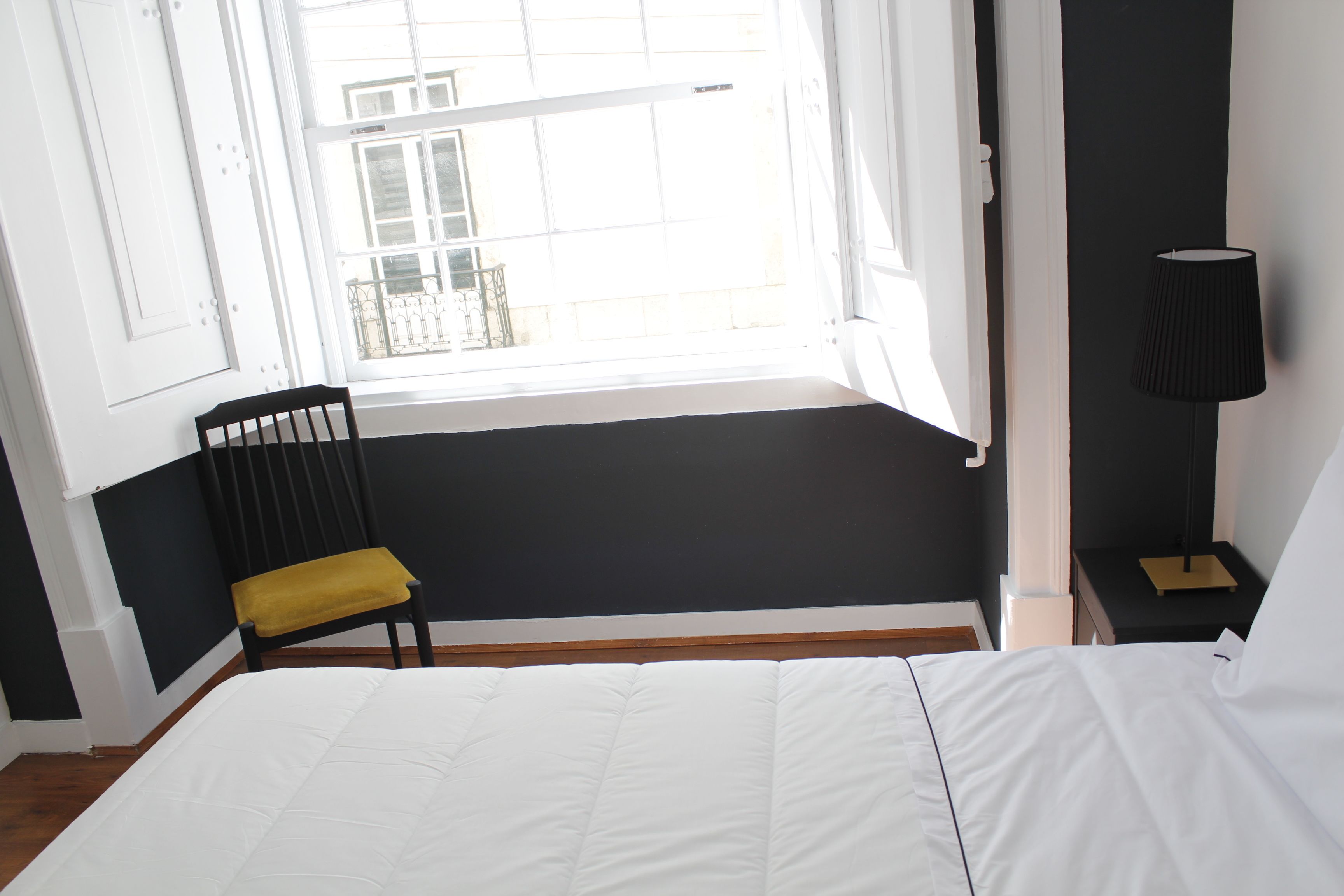 A bedroom with a large window, a bed, a chair, and a bedside table with a lamp.