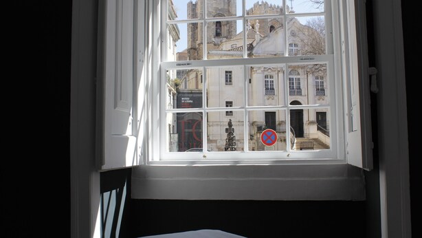 A window with a view of a historic building with multiple windows and a statue. - Largo da Sé - HOrigem (Lisbon)