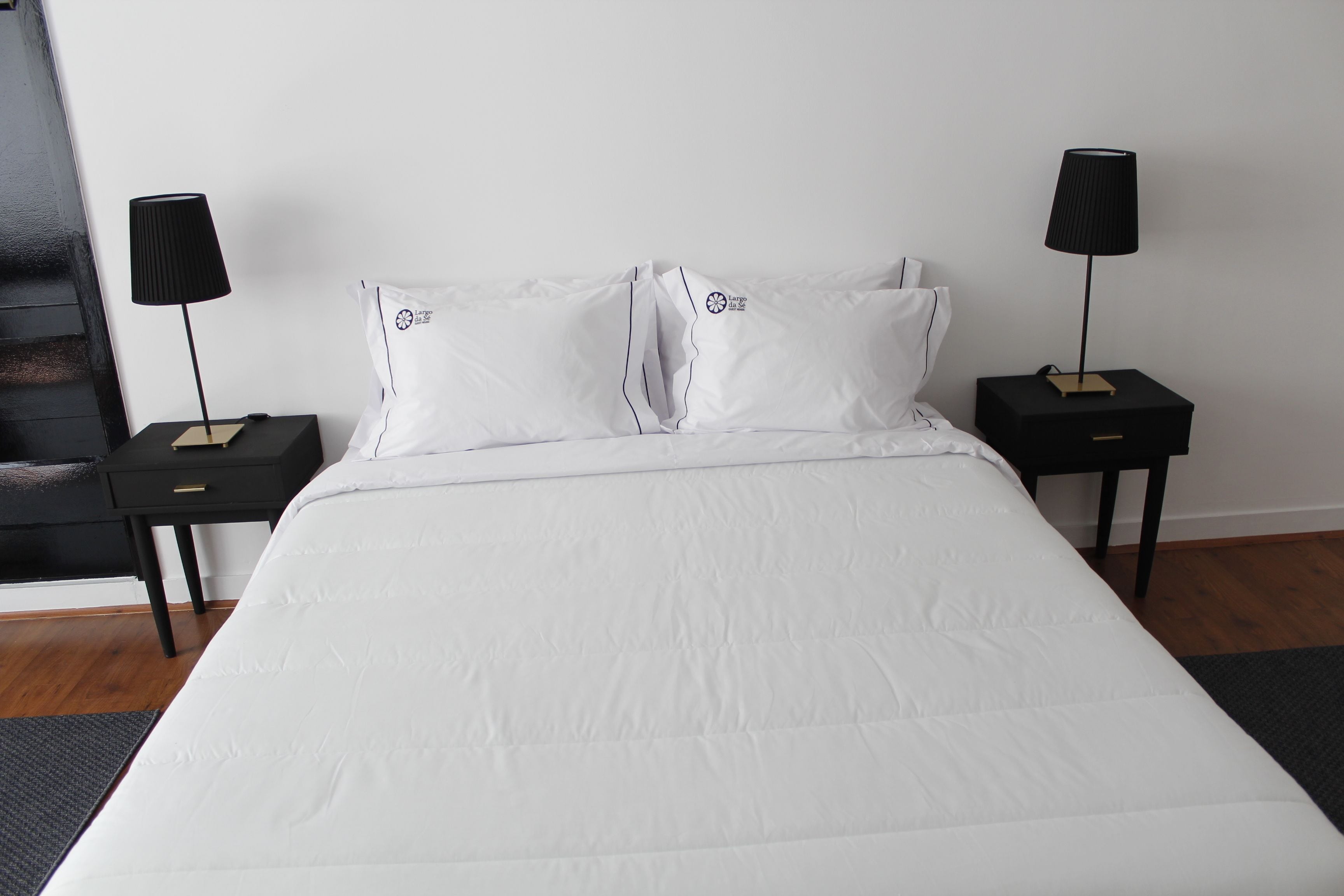 Een net opgemaakt bed met witte beddengoed, twee zwarte nachtkastjes met lampen, en een donker vloerkleed.