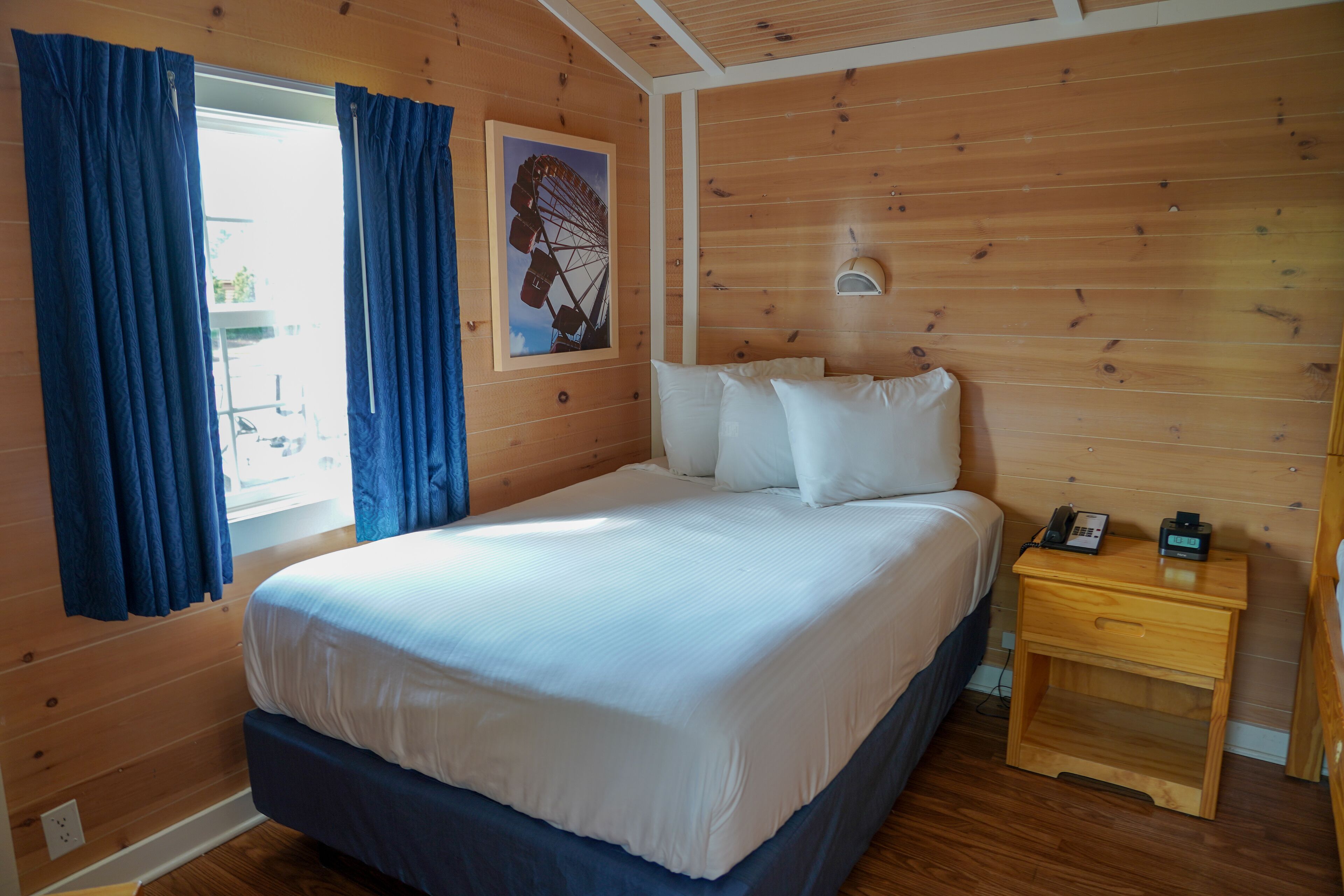 A room with a wooden floor, a bed with white bedding, a nightstand with a phone and a lamp, and a window with blue curtains.