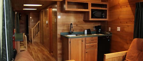 A compact kitchen with wooden cabinets, a microwave, and a coffee maker. There is a wooden staircase leading to an upper level.