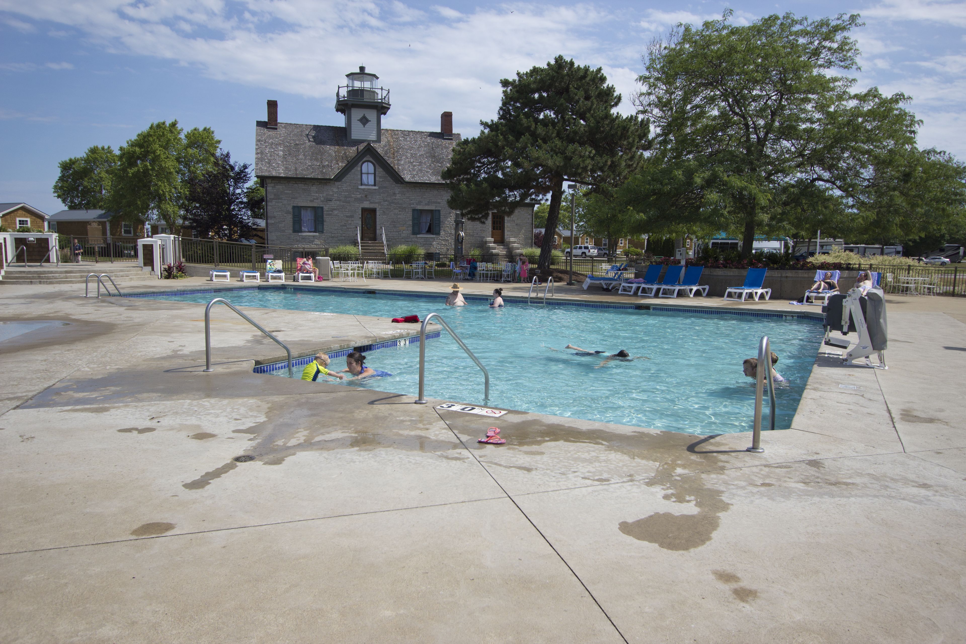 A swimming pool with a stone building in the background, a fence around the pool, and a few people swimming.