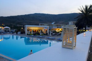 Una piscina con sillas de descanso, un farol blanco y un edificio con mesas al aire libre al fondo.