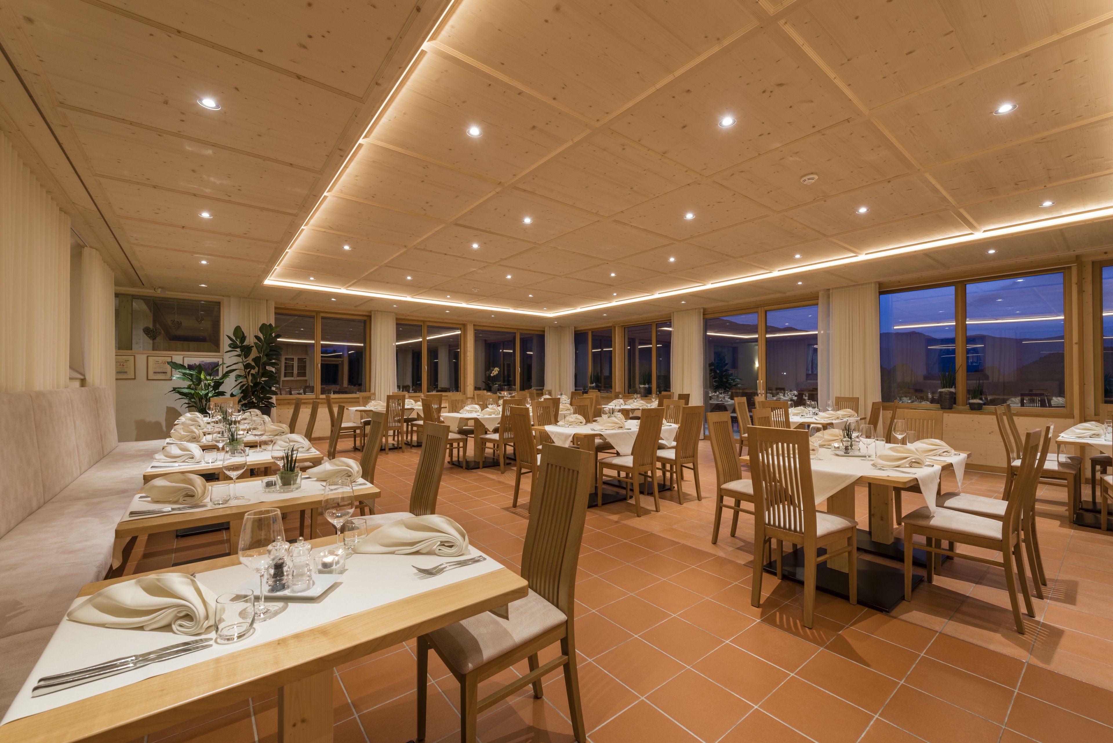 Een ruime eethoek met meerdere tafels en stoelen, grote ramen en een hoge plafond met inbouwverlichting.