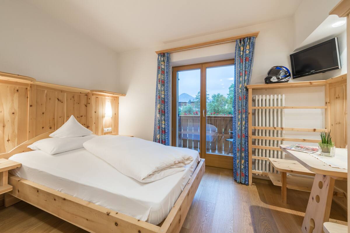 Un lit en bois avec du linge de lit blanc, un téléviseur posé sur une étagère en bois, et un balcon offrant une vue sur la verdure et les bâtiments.
