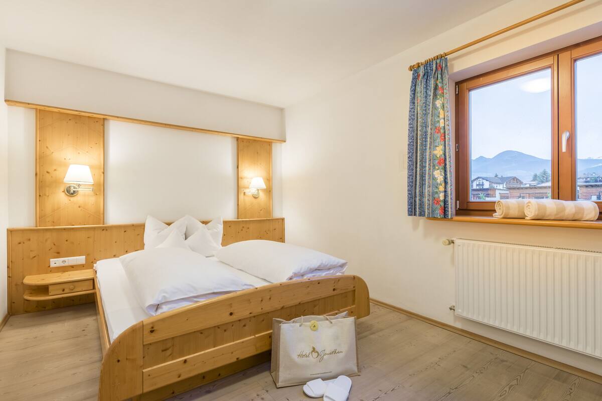Une chambre d’hôtel avec un lit en bois, deux lampes de chevet, une fenêtre avec des rideaux et une vue sur des immeubles et des montagnes.
