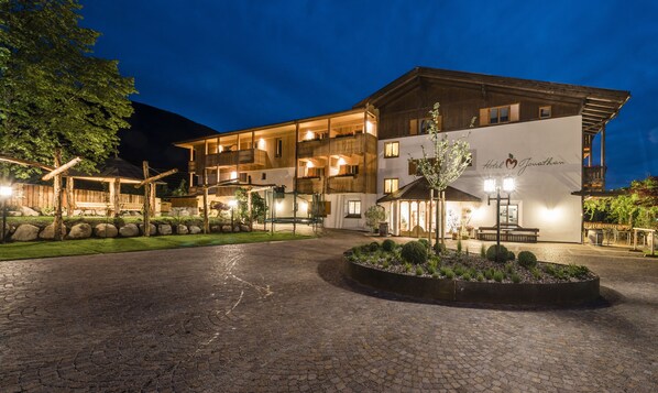 A multi-story hotel with a well-lit entrance, outdoor seating area, and a landscaped garden. - Hotel Jonathan (Naz Sciaves)