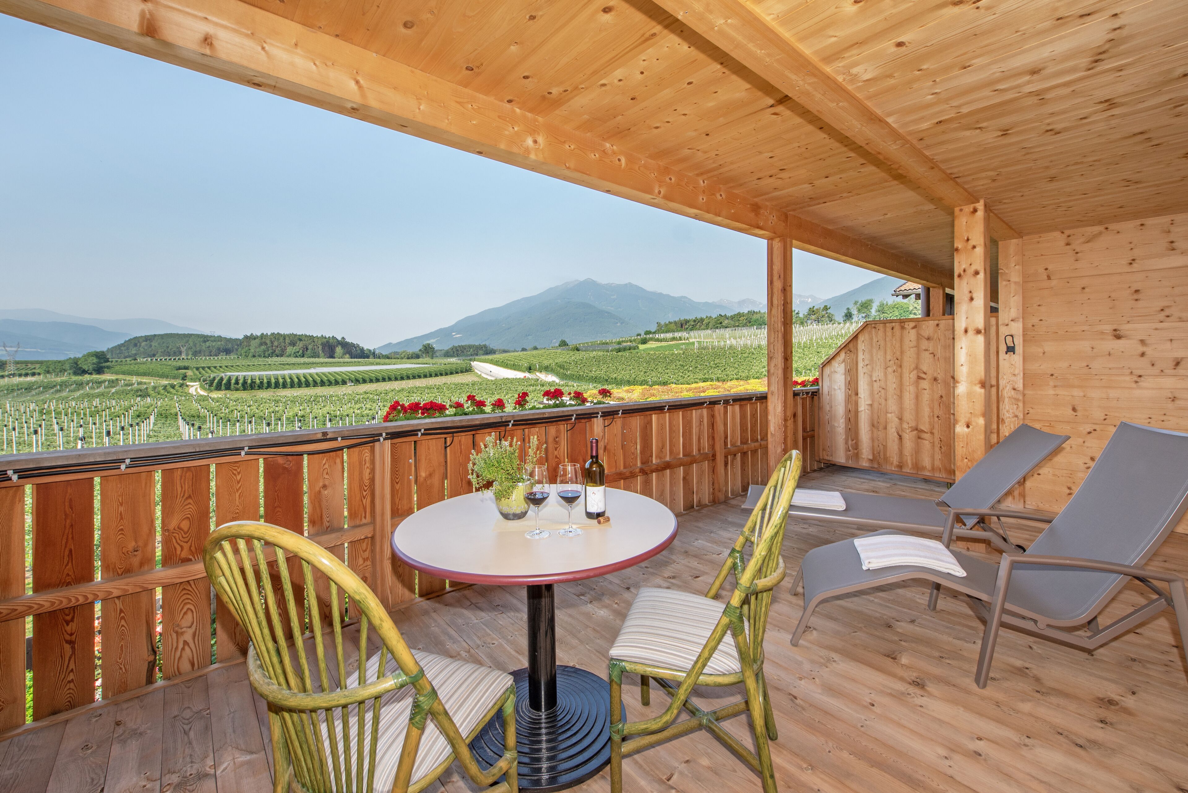 Eine Holzterrasse mit rundem Tisch und Stühlen, mit Blick auf einen Weinberg und Berge.