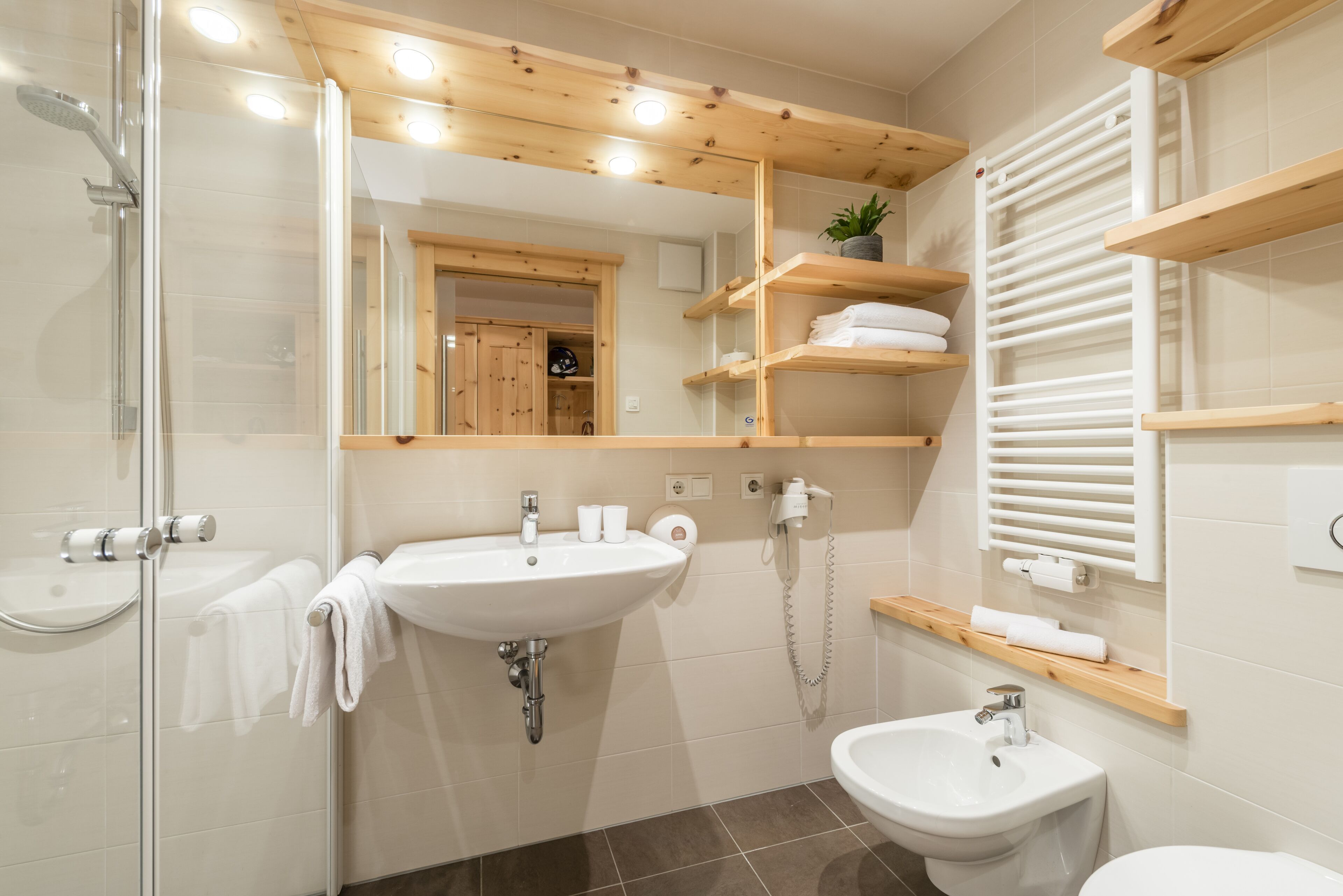 A modern bathroom with a glass shower enclosure, a sink, a bidet, and wooden shelves with towels.