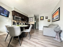 Una sala moderna con un sofá gris, un comedor con una mesa para cuatro, y una cocina con armarios de madera oscura y electrodomésticos de acero inoxidable.