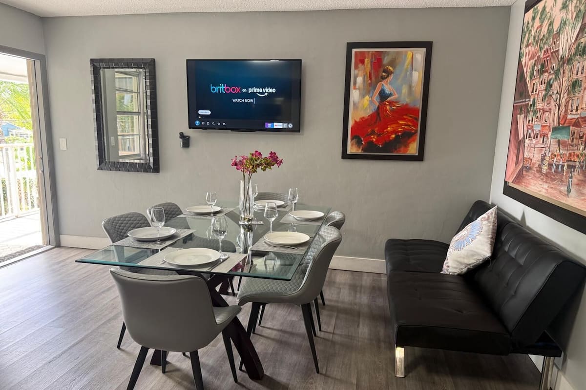 Een moderne eetruimte met een glazen tafel voor zes personen, een zwarte leren bank en een flatscreen televisie waarop 'Britbox on Prime Video' wordt weergegeven.