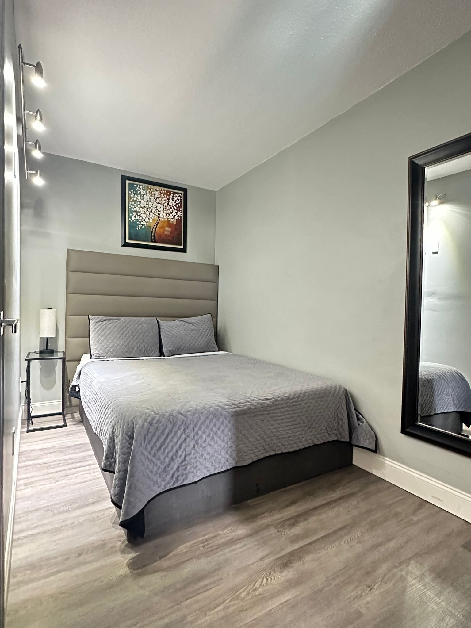 A bedroom with a bed, a bedside table, a lamp, a framed artwork, and a large mirror.