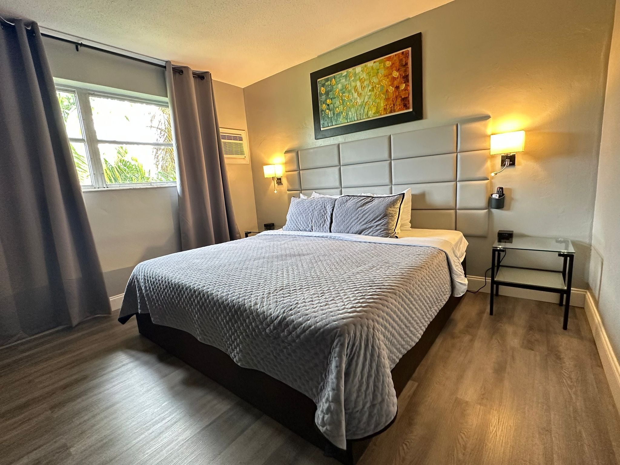 Quarto de hotel moderno com uma cama grande, duas luminárias de cabeceira, um quadro sobre a cama e uma mesinha com uma luminária.