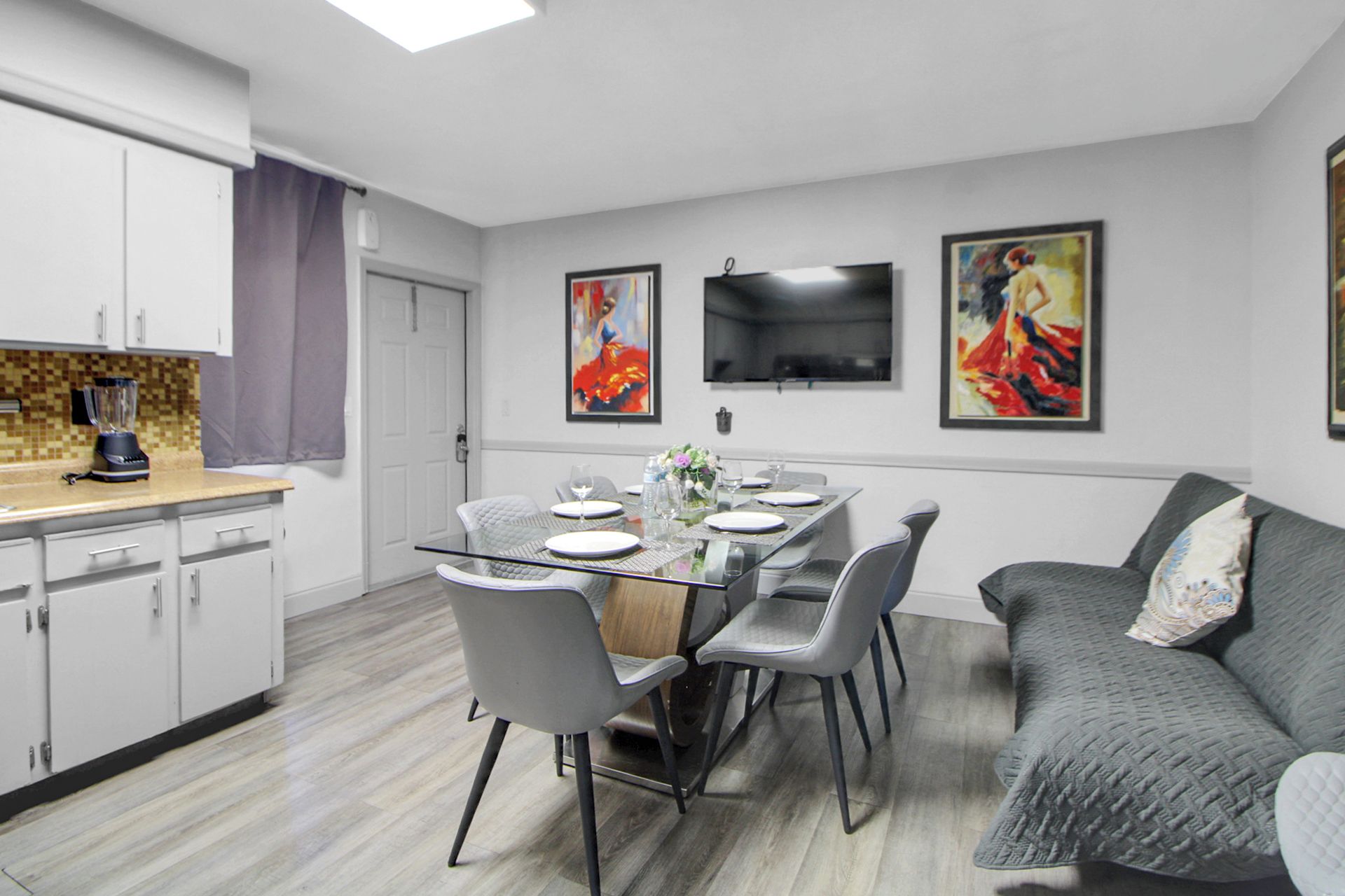 Una cocina moderna con armarios blancos, una mesa de comedor para cuatro personas y un sofá gris con un cojín.