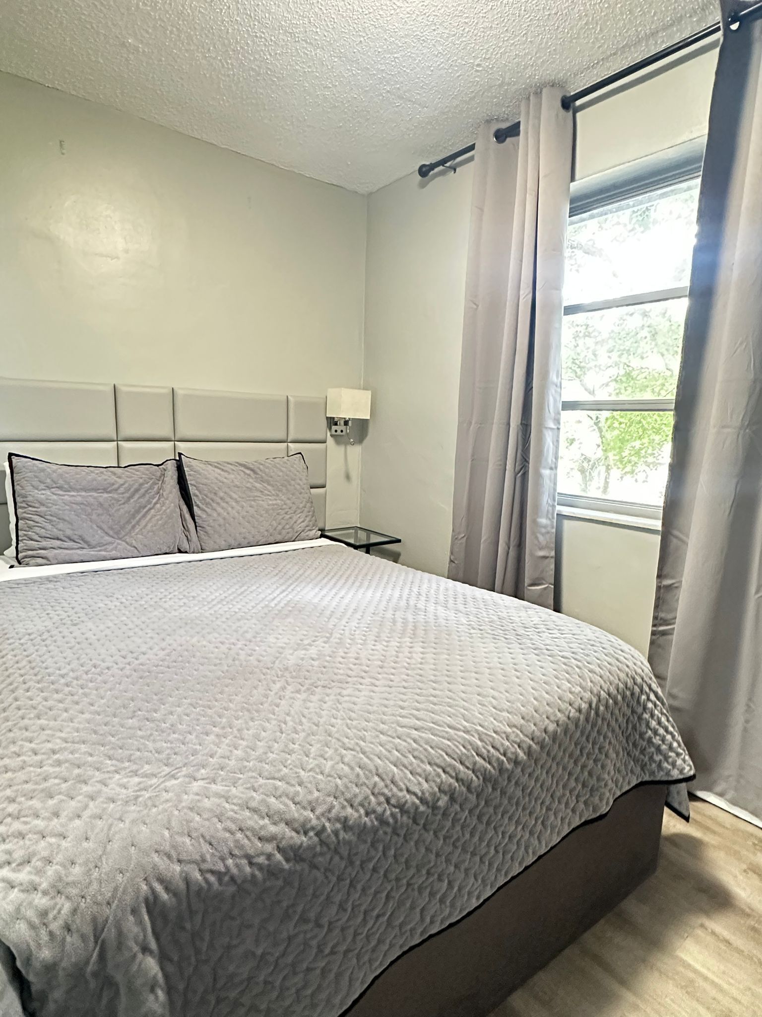 Un dormitorio con una cama grande, una mesita de noche, una lámpara de pared y una ventana con cortinas.