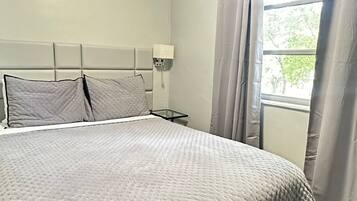Un dormitorio con una cama grande, una mesita de noche, una lámpara de pared y una ventana con cortinas.