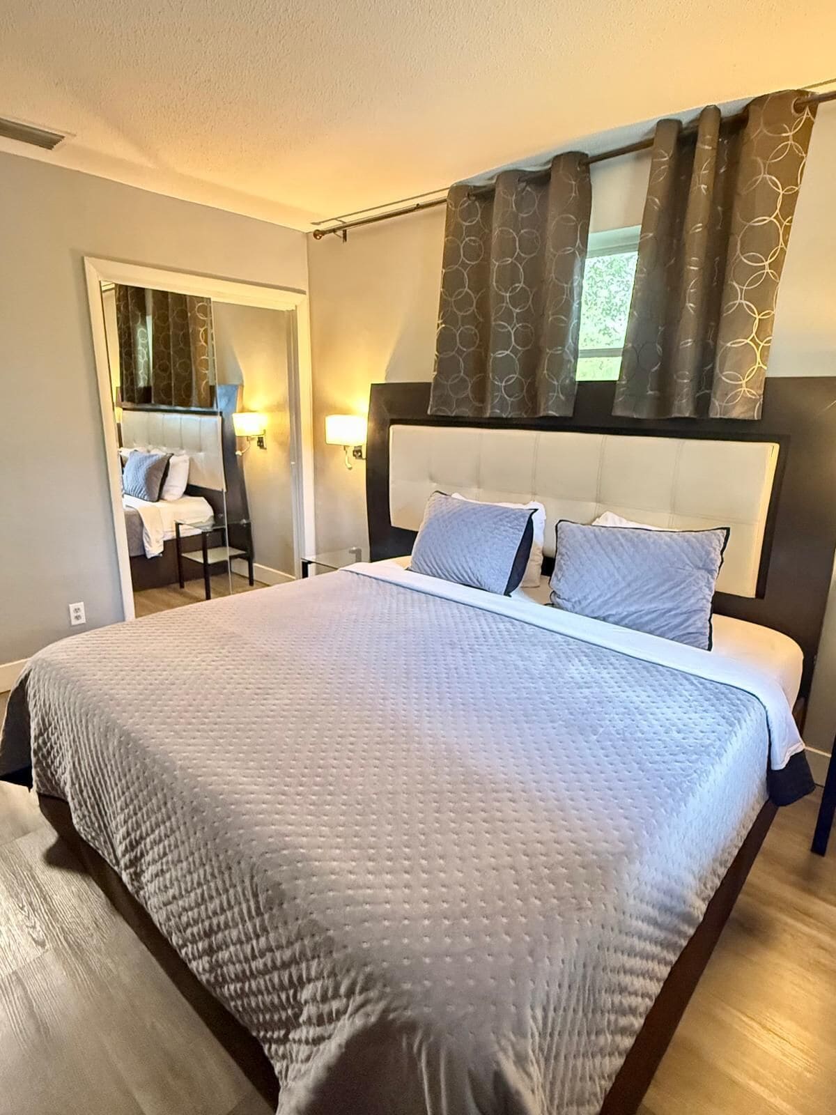 Quarto de hotel moderno com uma cama grande, cabeceira com espelho e vista para o exterior pela janela.