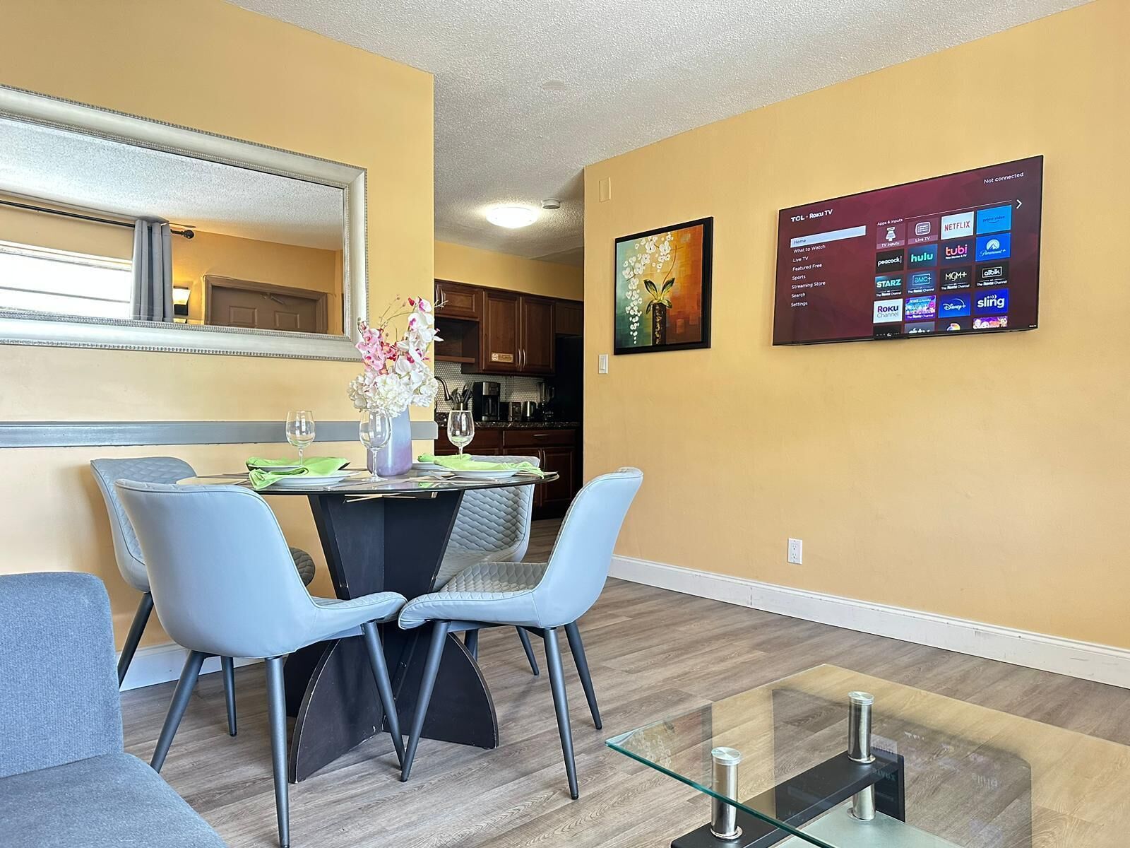 Un salon moderne avec un coin repas, comprenant une table basse en verre, deux fauteuils bleus, un vase avec des fleurs et un téléviseur fixé au mur qui diffuse différents services de streaming.