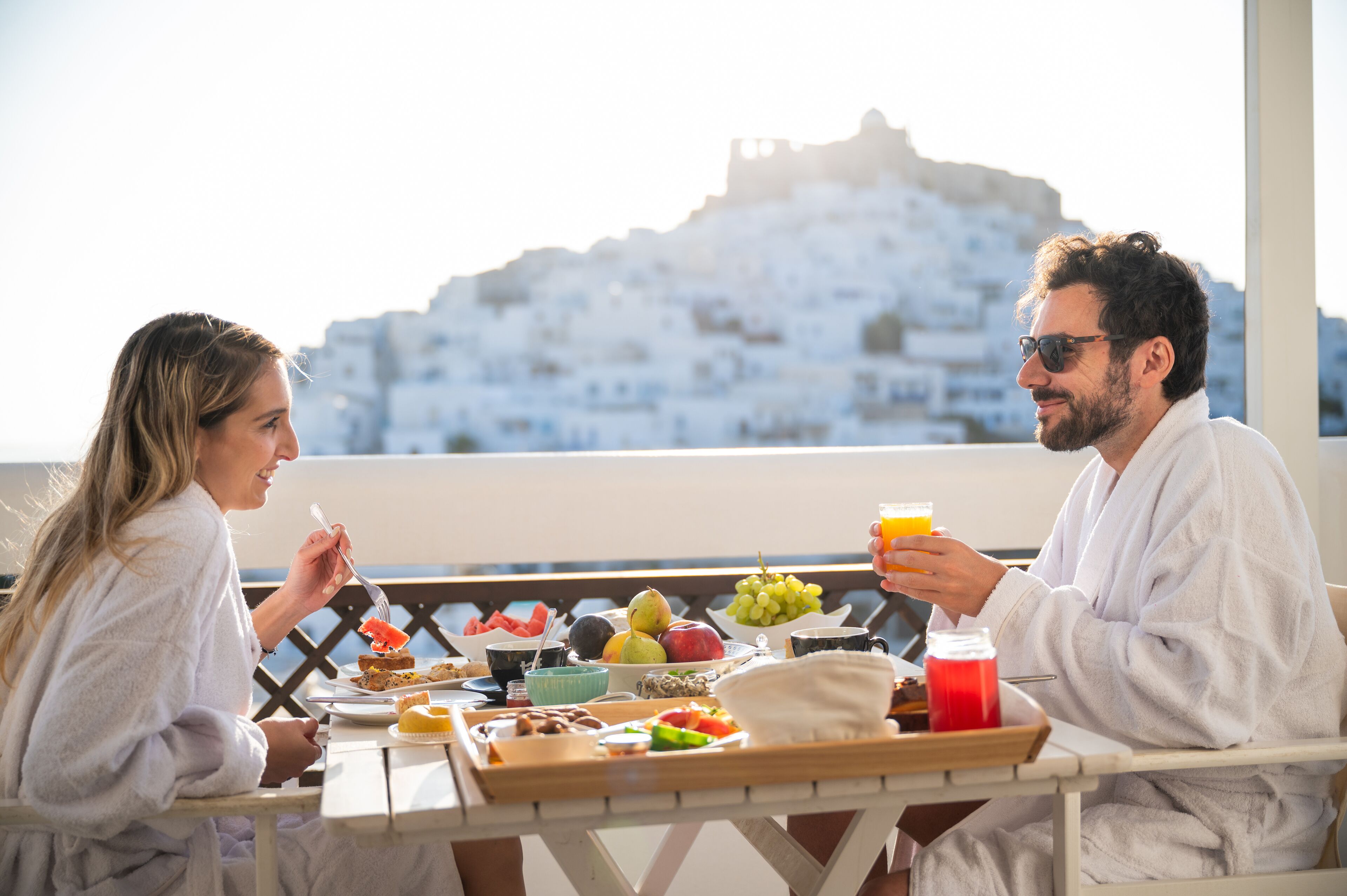 A couple dining on a balcony with a scenic view of a white-washed building.