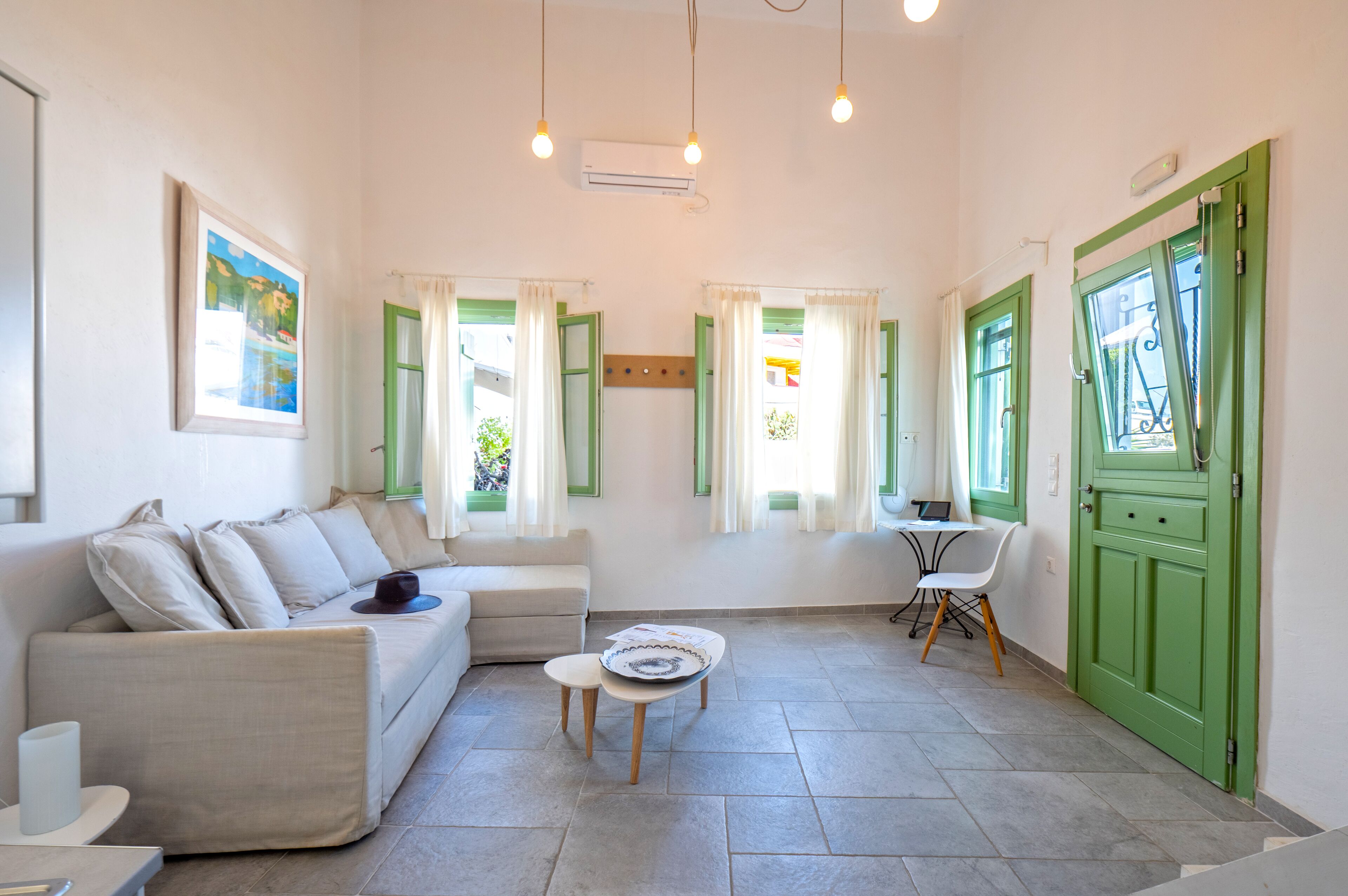 Una sala de estar con un sofá, una mesita y un sillón. Hay dos ventanas con contraventanas verdes y cortinas blancas.
